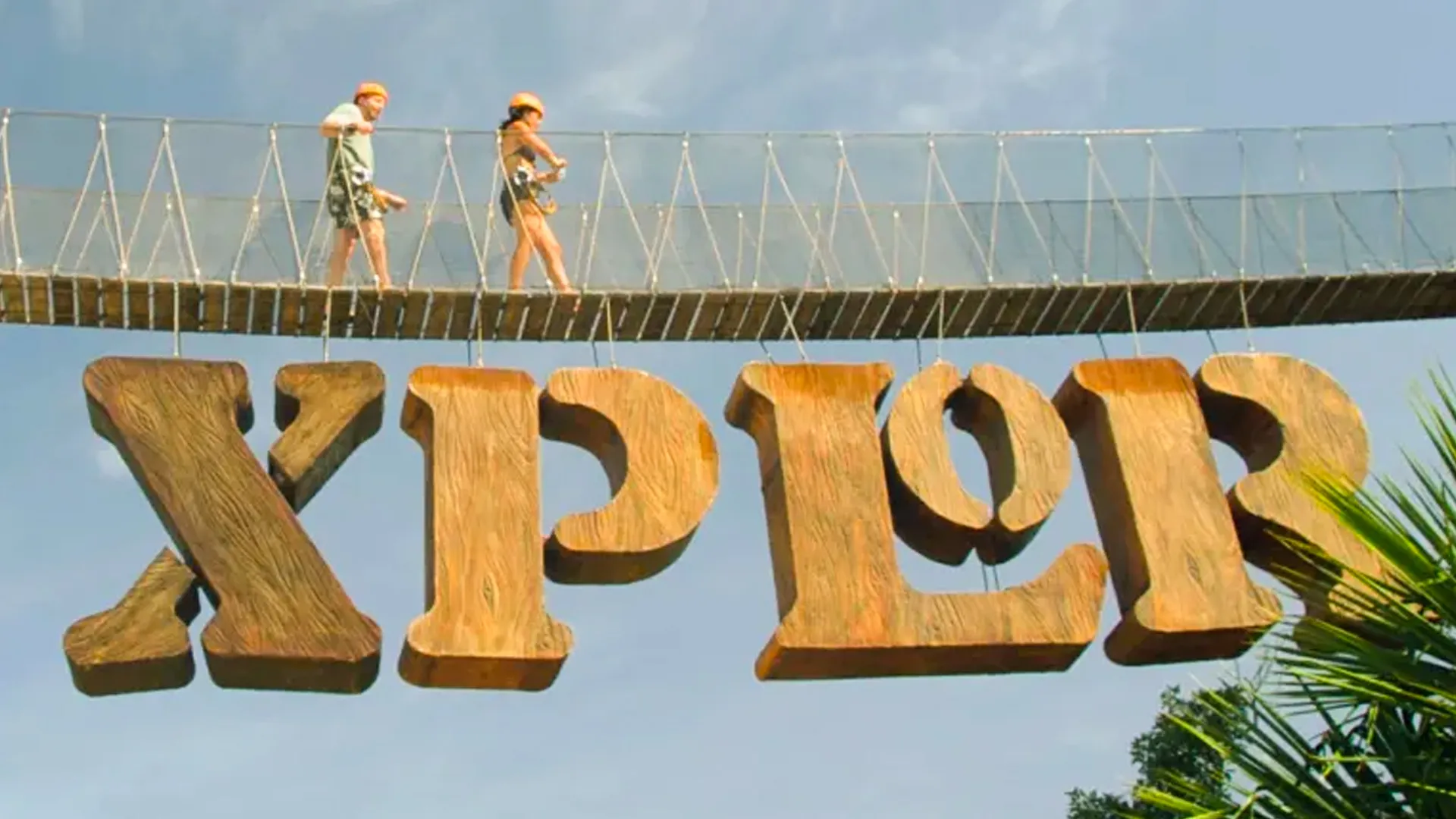 Two people walking across a rope bridge with a large "XPLOR" sign below, set against a clear blue sky and tropical greenery.