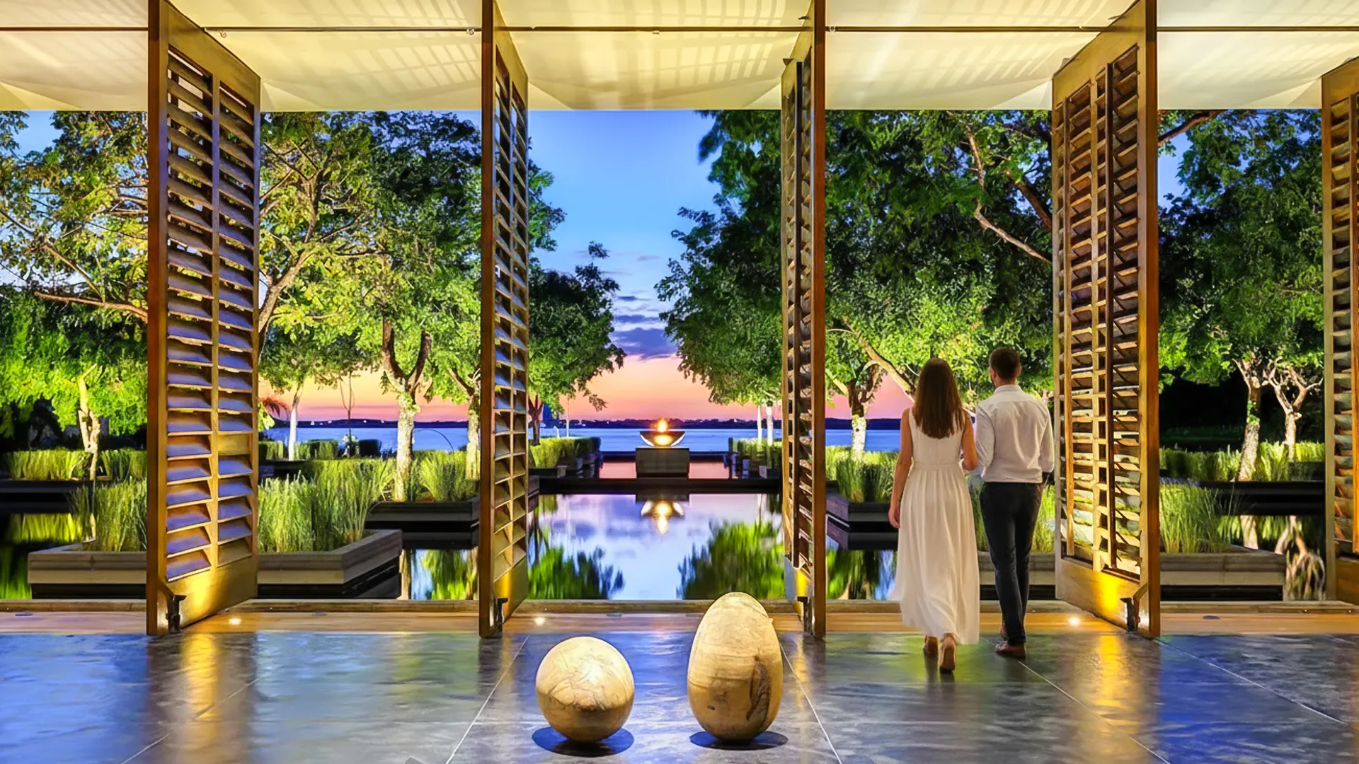 Couple walking by lantern-lit wooden deck at Nizuc Resort Cancun during sunset, surrounded by tropical gardens.