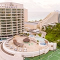 Aerial view of the rooftop pool area at Seadust Cancun Family Resort showing a circular pool with a central island, surrounded by lounge chairs, thatched umbrellas, and a multi-story resort building with balconies, set against lush greenery and a cloudy sky.