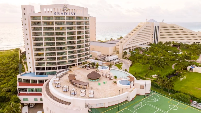 Aerial view of the rooftop pool area at Seadust Cancun Family Resort showing a circular pool with a central island, surrounded by lounge chairs, thatched umbrellas, and a multi-story resort building with balconies, set against lush greenery and a cloudy sky.