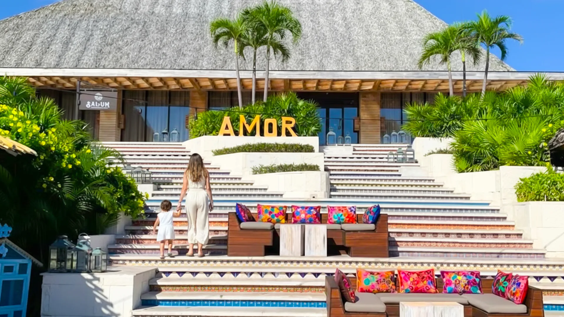 Entrance of Salum Beach Club at Vidanta with AMOR sign, colorful cushions, palm trees, and a mother walking with her child.