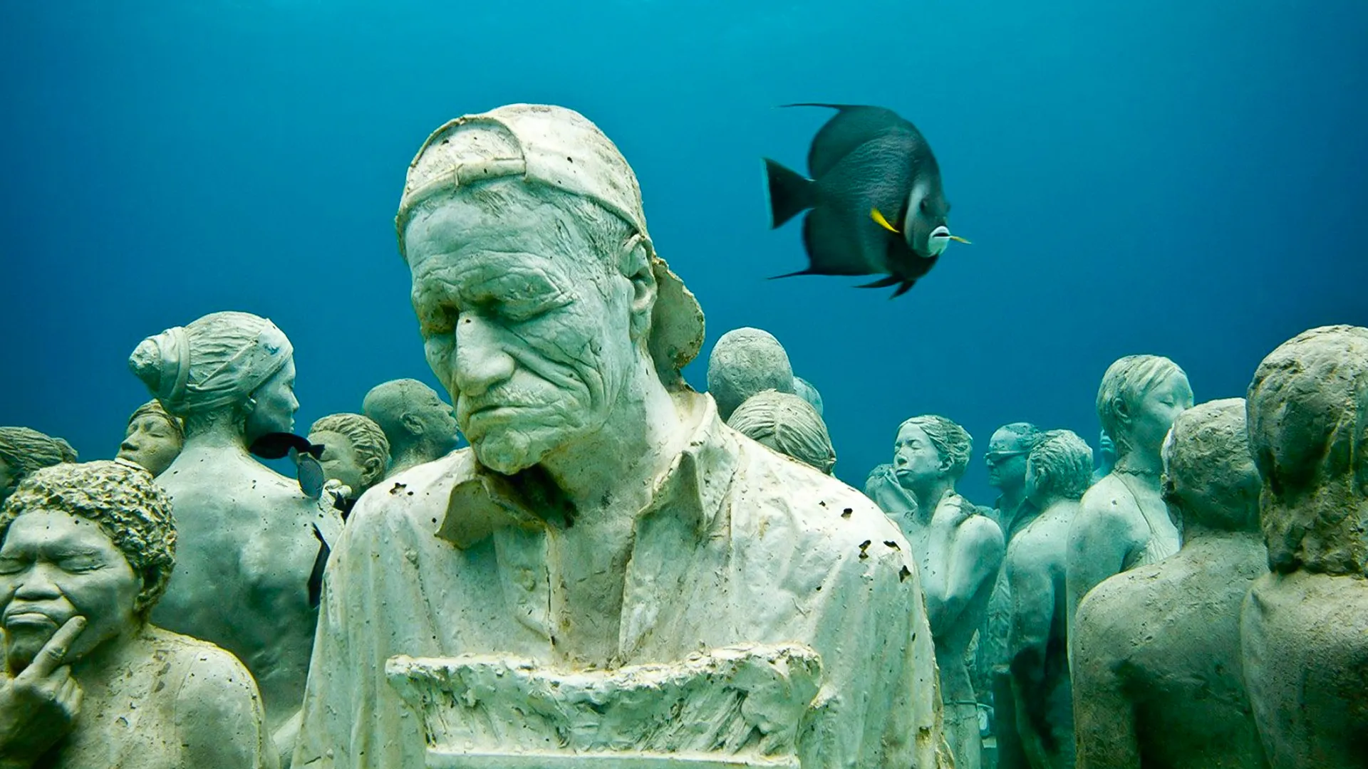 An underwater scene featuring a statue of a woman with a thoughtful expression, surrounded by numerous other statues and a black fish swimming nearby.