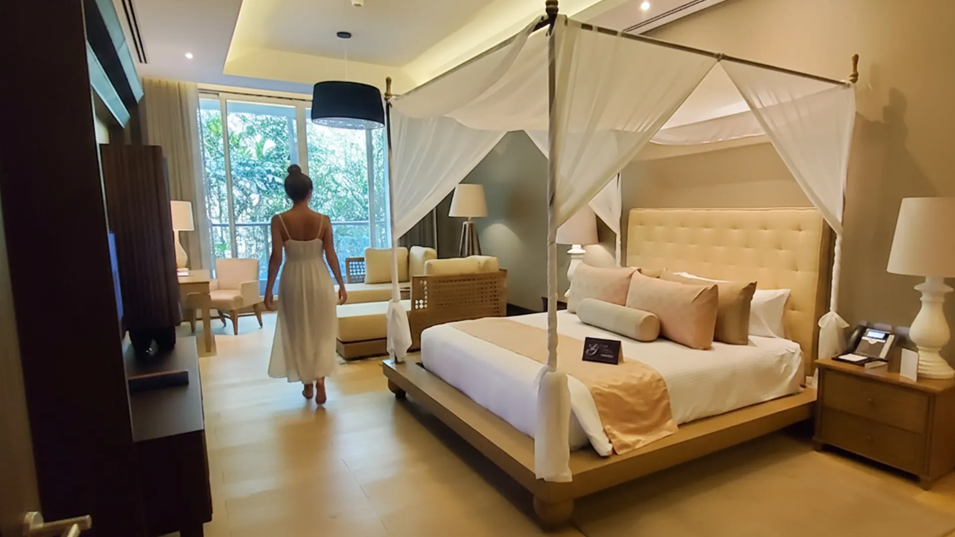 A luxurious hotel room with a large canopy bed featuring white drapes, beige bedding with pink and peach pillows, a person in a white dress standing near the bed, a sitting area with beige chairs, and large windows with a view of greenery outside.