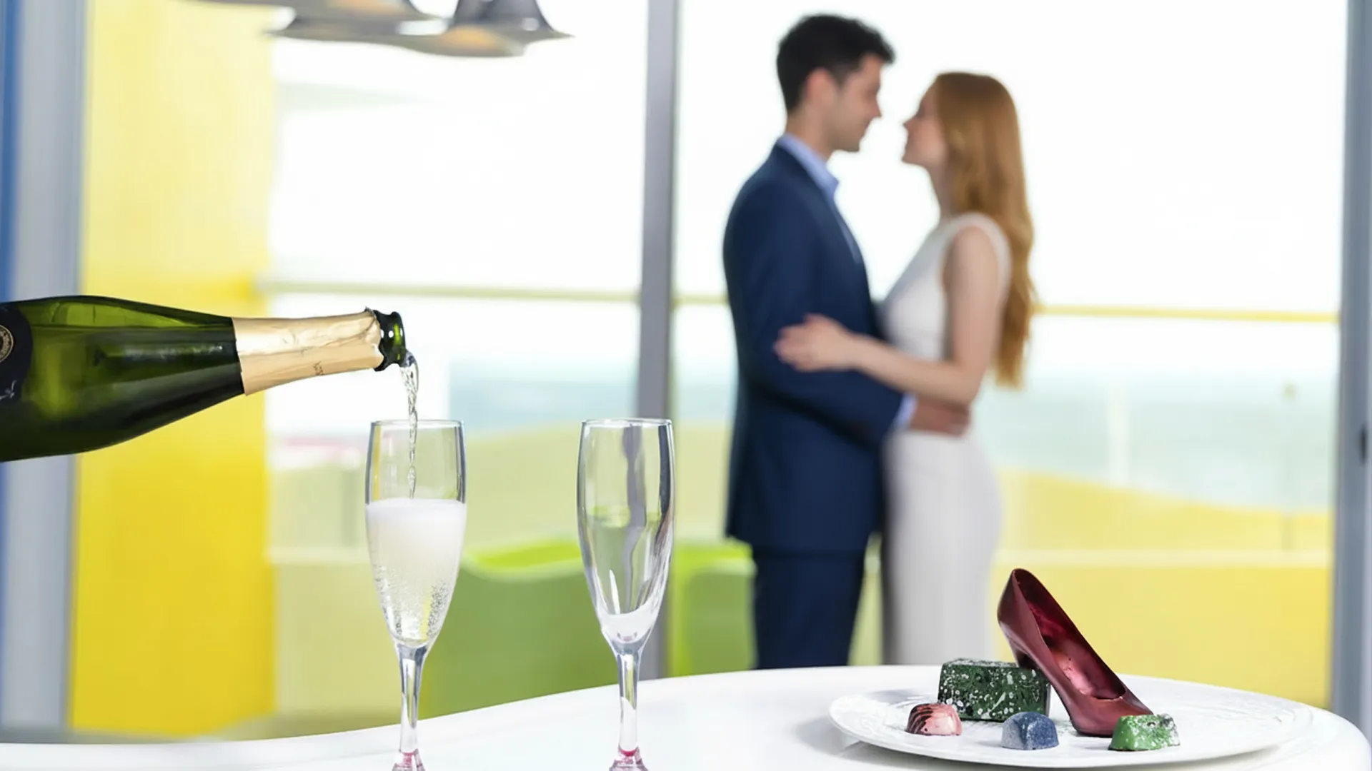 A couple in formal attire embracing near a window with a sea view, while a bottle of champagne is poured into two glasses on a table with chocolates and a high-heeled shoe-shaped decoration.