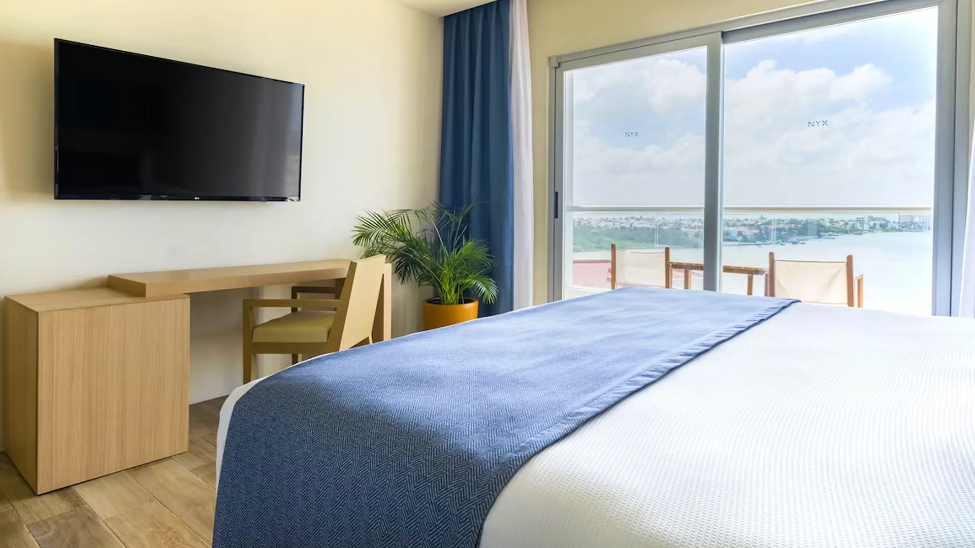 A modern hotel room with a neatly made bed, a flat-screen TV on the wall, a wooden desk with a chair, a potted plant, and a large glass door opening to a balcony with chairs and a view of the ocean.