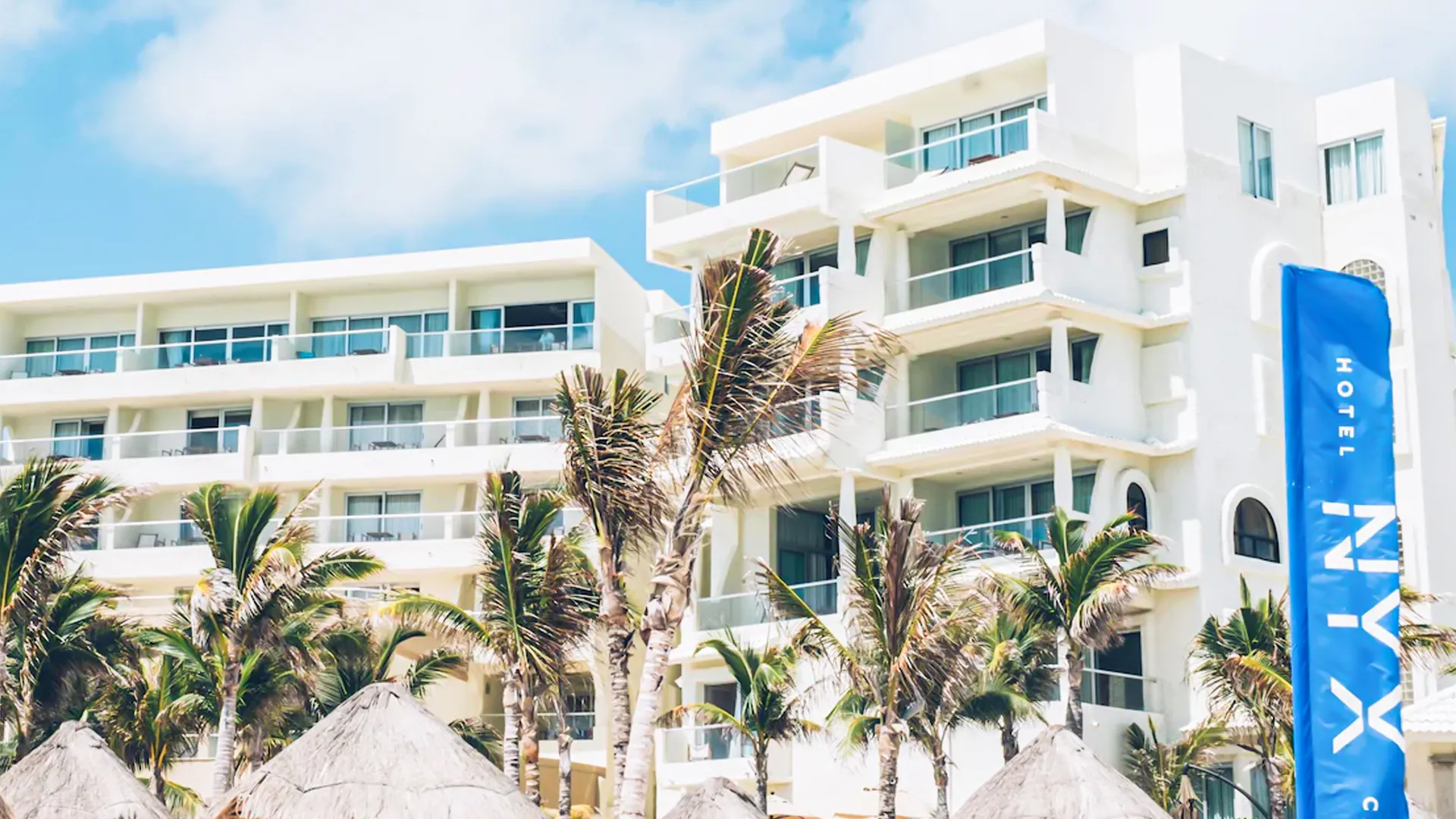 A multi-story white hotel building with balconies, surrounded by palm trees and thatched-roof structures, with a blue flag displaying "NYX Hotel" in front.