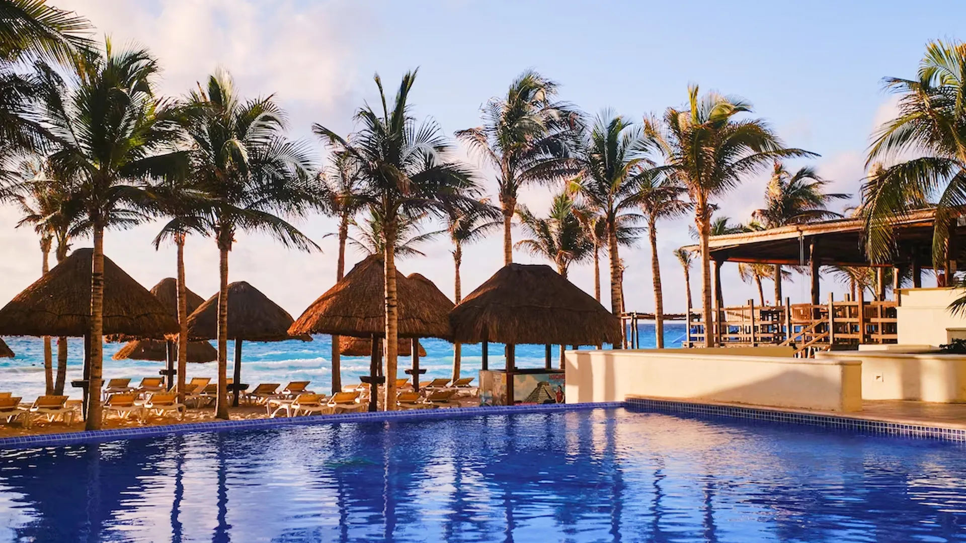 A tropical resort pool with clear blue water, surrounded by palm trees and thatched-roof umbrellas, with lounge chairs and the ocean visible in the background under a partly cloudy sky.