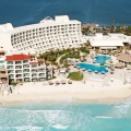 Aerial view of Secrets Playa Mujeres resort in Cancun, featuring a large white hotel building, multiple swimming pools, a sandy beach, and turquoise ocean waters with tropical landscaping.