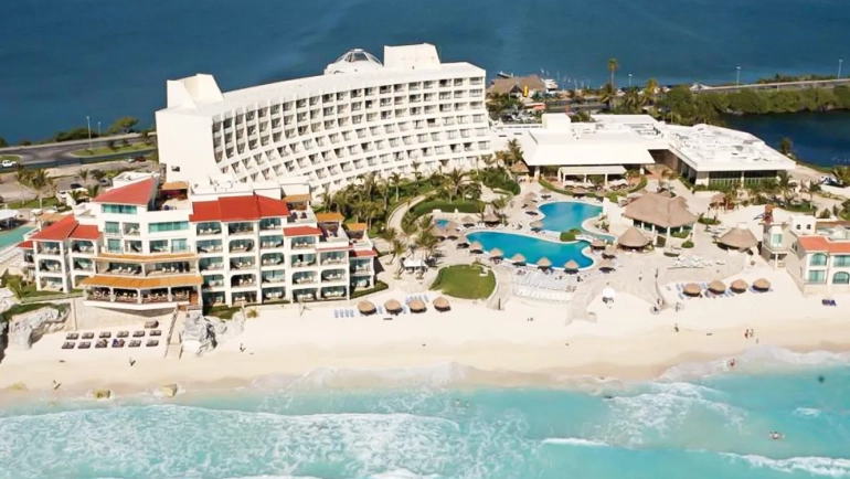 Aerial view of Secrets Playa Mujeres resort in Cancun, featuring a large white hotel building, multiple swimming pools, a sandy beach, and turquoise ocean waters with tropical landscaping.