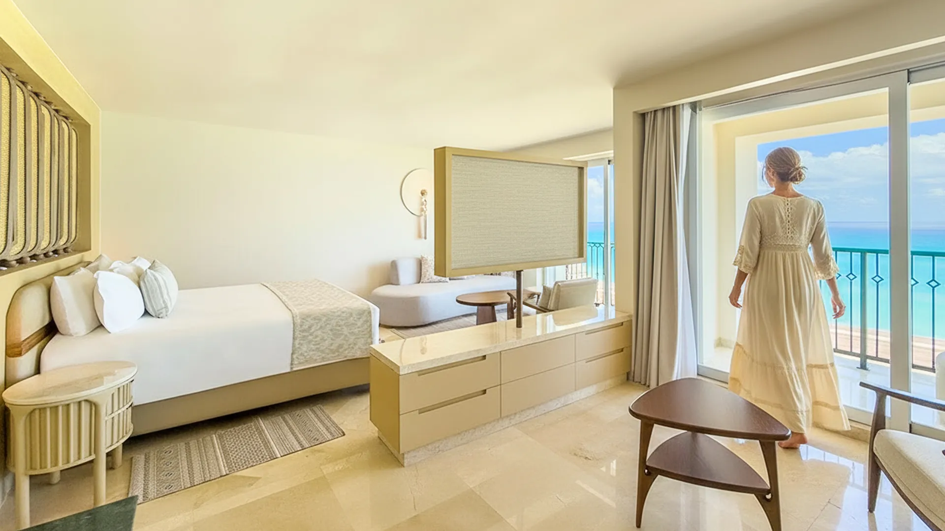 A woman in a white dress standing by a large window with an ocean view, inside a modern hotel room with a bed, sofa, and wooden furniture.