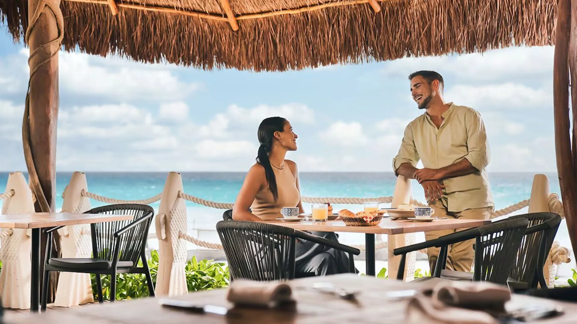 A couple enjoying breakfast at an outdoor table under a thatched roof, with a view of the turquoise ocean and beach in the background.