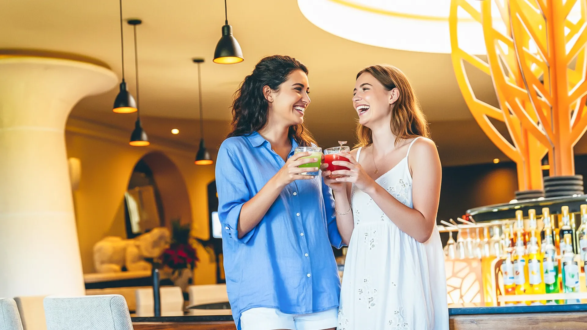 Two women laughing and toasting with colorful drinks at a modern bar with warm lighting and a decorative orange tree sculpture.