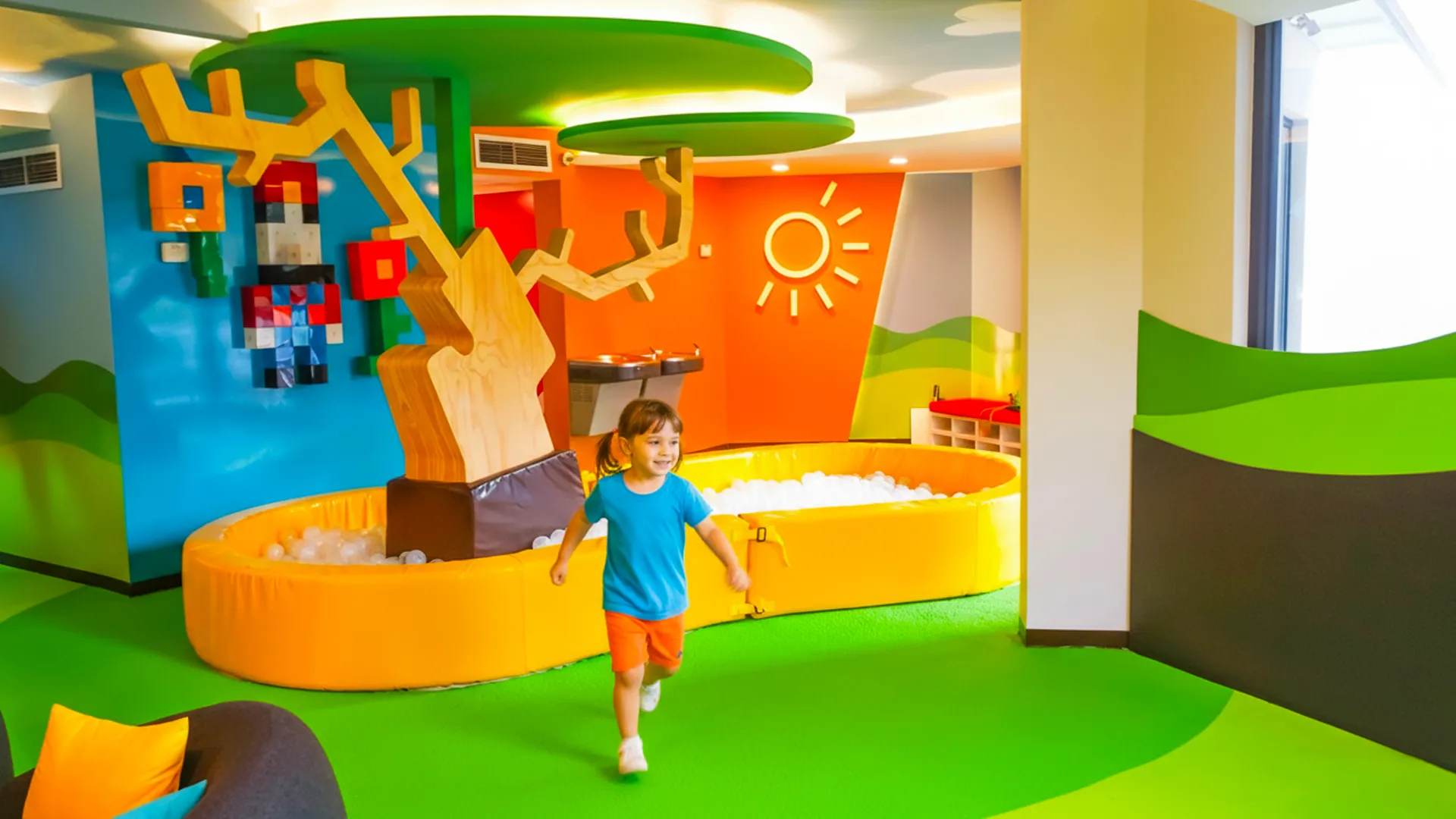 A child running joyfully in a colorful indoor play area with a ball pit, a large tree sculpture, and vibrant wall decorations.