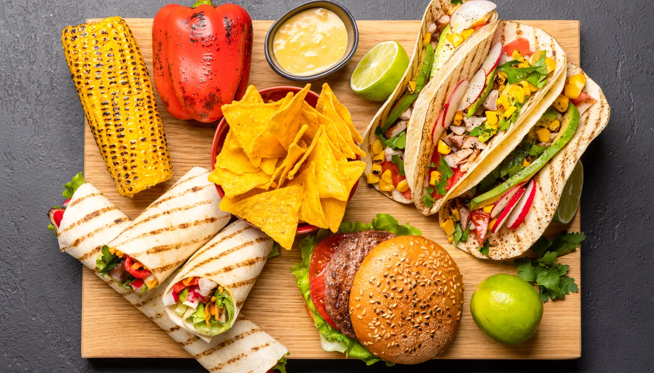 A wooden board with a variety of foods including tacos, wraps, a burger, corn on the cob, tortilla chips, grilled vegetables, and lime wedges.