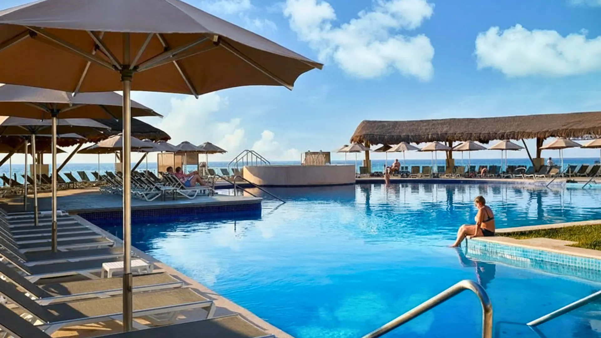 A large outdoor swimming pool with clear blue water, surrounded by lounge chairs, umbrellas, and a thatched-roof structure, with the ocean in the distance.