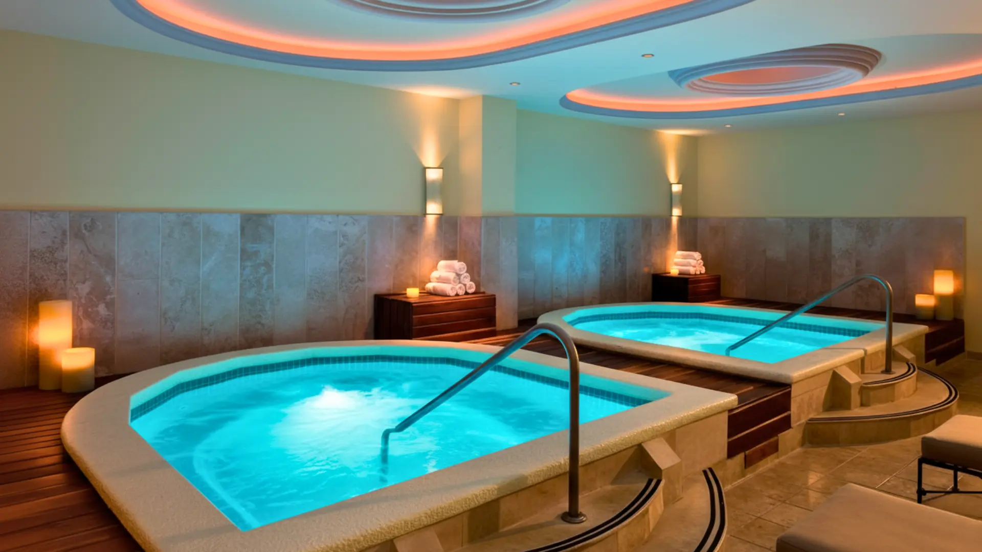 A serene indoor spa area featuring two glowing hot tubs with handrails, stone walls, wooden accents, and soft lighting from candles and ceiling fixtures.
