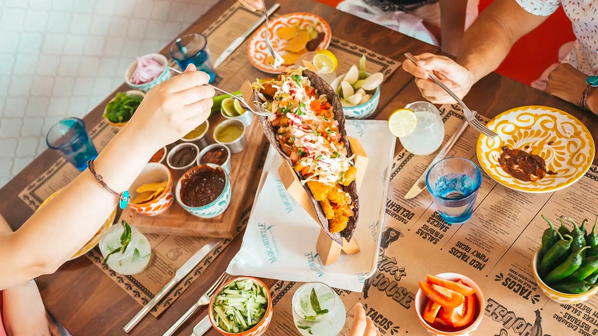 Overhead view of guests sharing tacos and cocktails at a vibrant Margaritaville Cancun restaurant table filled with colorful dishes and sauces.