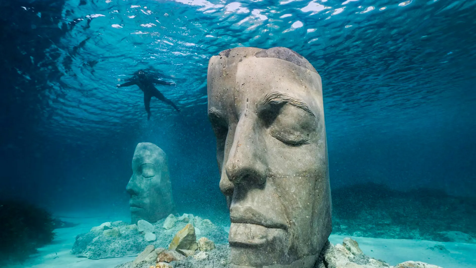 An underwater view of a large stone head statue with a solemn face, with a scuba diver swimming in the background and a second statue visible.