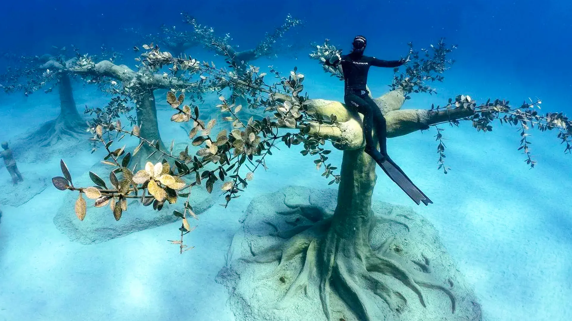 A scuba diver sitting on an underwater sculpture resembling a tree with coral-like branches, set against a sandy ocean floor.