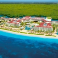 An aerial view of a Cancun resort along a sandy beach, surrounded by lush green mangroves and turquoise waters, with multiple resort buildings featuring red-tiled roofs under a clear sky.