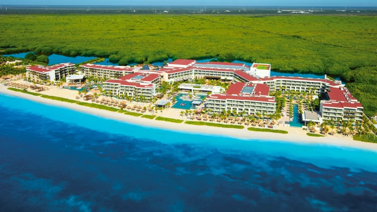 An aerial view of a Cancun resort along a sandy beach, surrounded by lush green mangroves and turquoise waters, with multiple resort buildings featuring red-tiled roofs under a clear sky.
