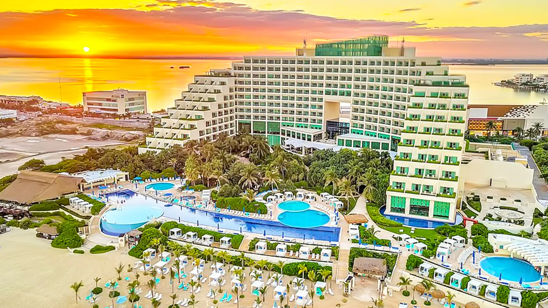 Aerial view of an unnamed resort with a multi-story building, multiple blue pools, white lounge chairs with umbrellas, green lawns, and palm trees against a sunset sky.