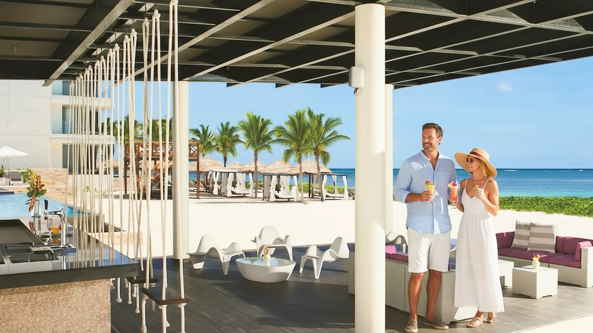A couple enjoying tropical cocktails at a stylish beachfront lounge at Breathless Cancun Soul, overlooking palm trees and the turquoise Caribbean Sea.