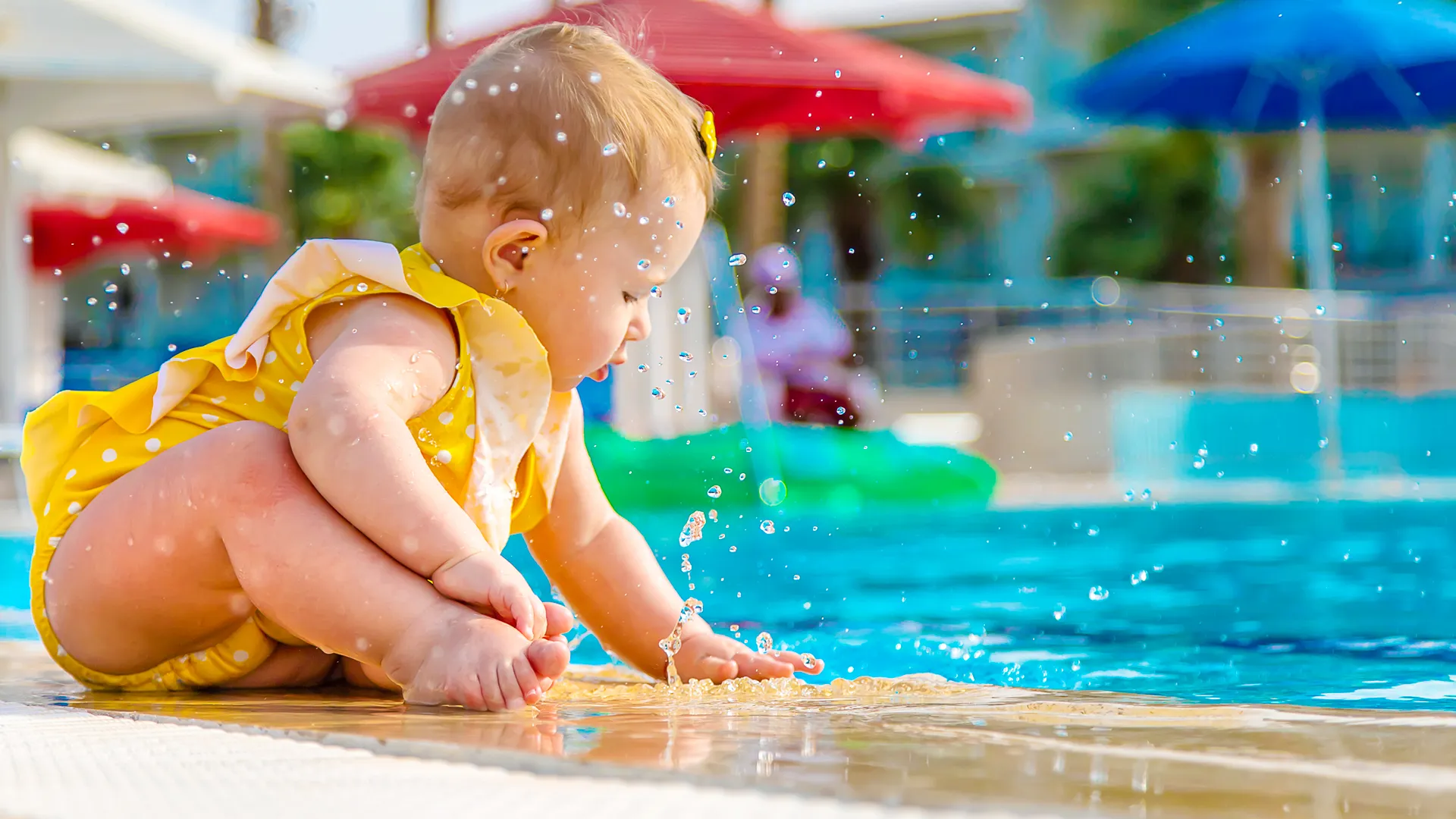 A young child in a yellow polka-dot swimsuit splashing water at the edge of a blue pool at Ventura Park, with colorful umbrellas and blurred background elements under a bright sky.