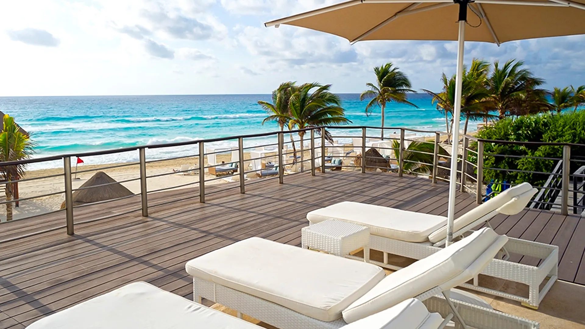 A wooden terrace with white lounge chairs and umbrellas, overlooking a sandy beach and turquoise ocean under a partly cloudy sky.