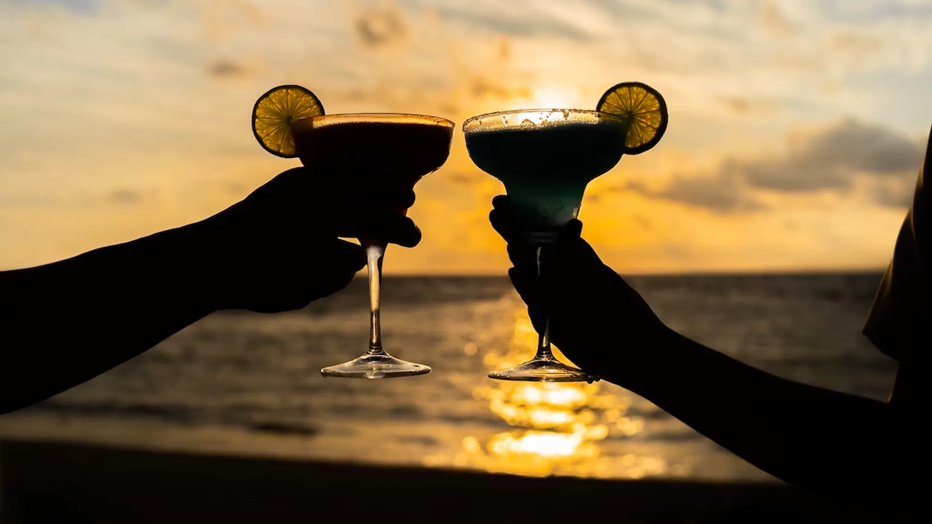 Silhouette of two guests toasting with margarita glasses at Margaritaville Island Reserve Riviera Cancun during sunset by the beach.