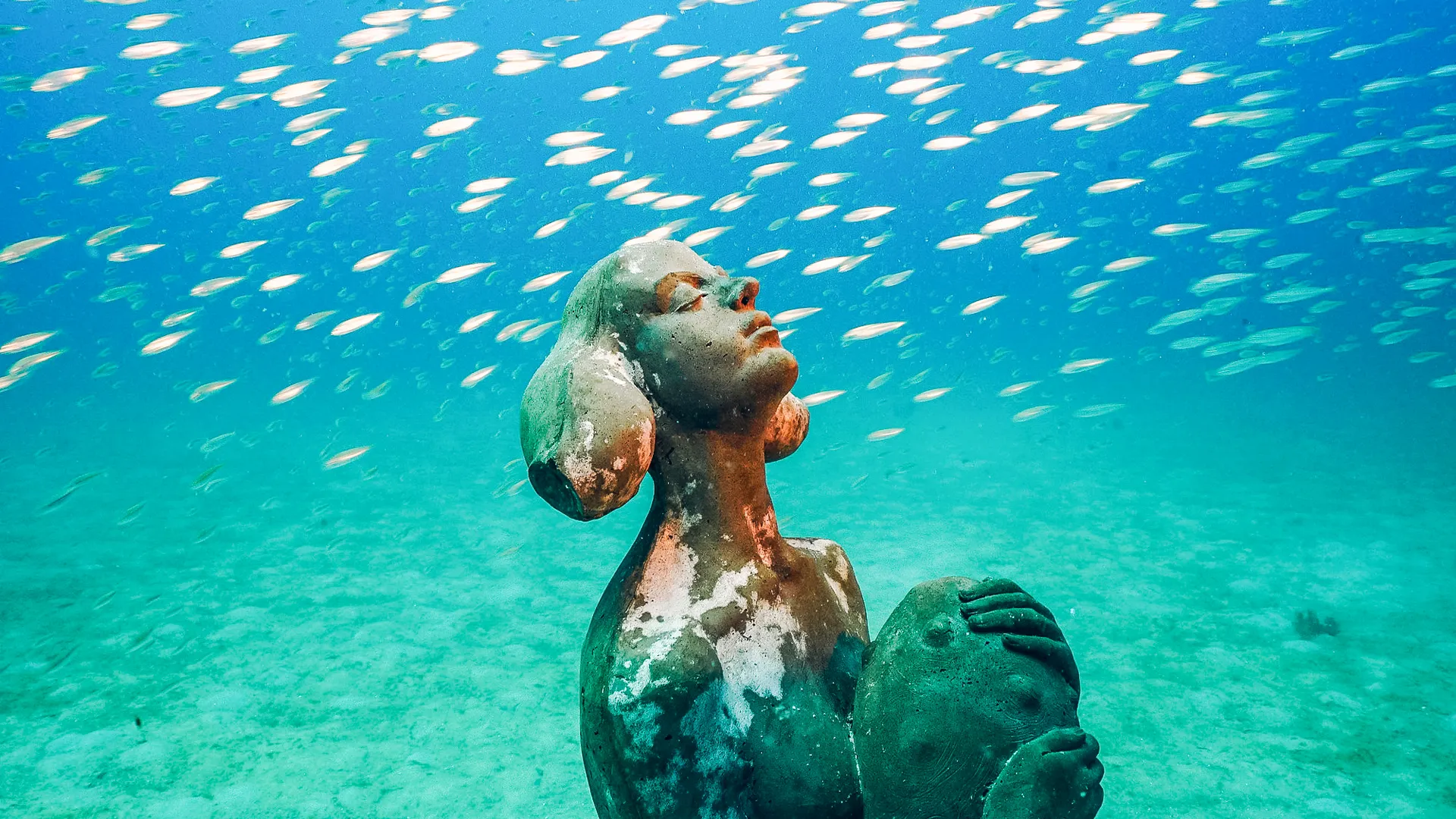 An underwater sculpture of a woman with her hands on her face, surrounded by a school of small fish in a vibrant blue ocean setting.
