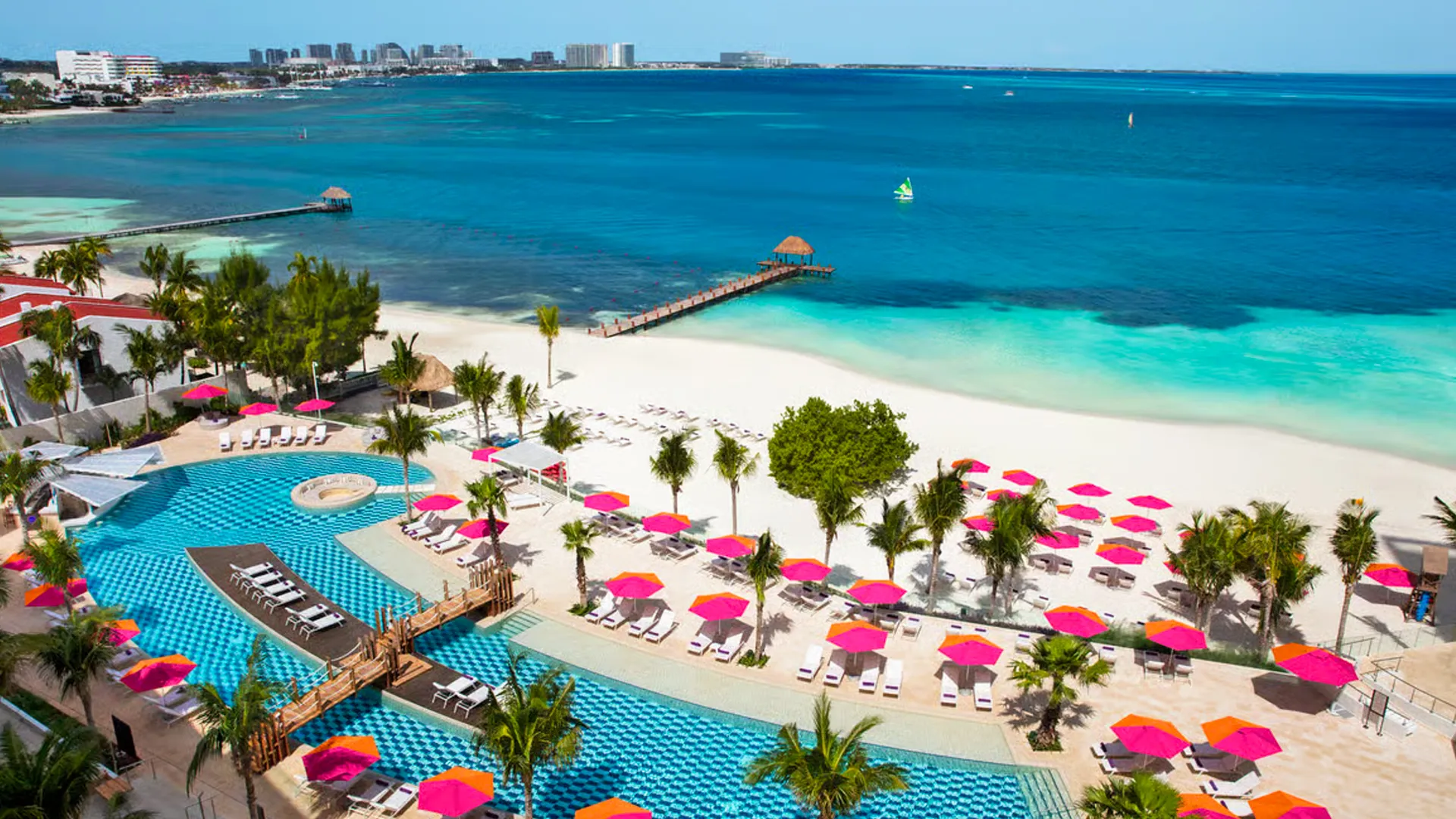Aerial view of Breathless Cancun Soul’s beach and infinity pool, featuring pink umbrellas, palm trees, and turquoise Caribbean waters.