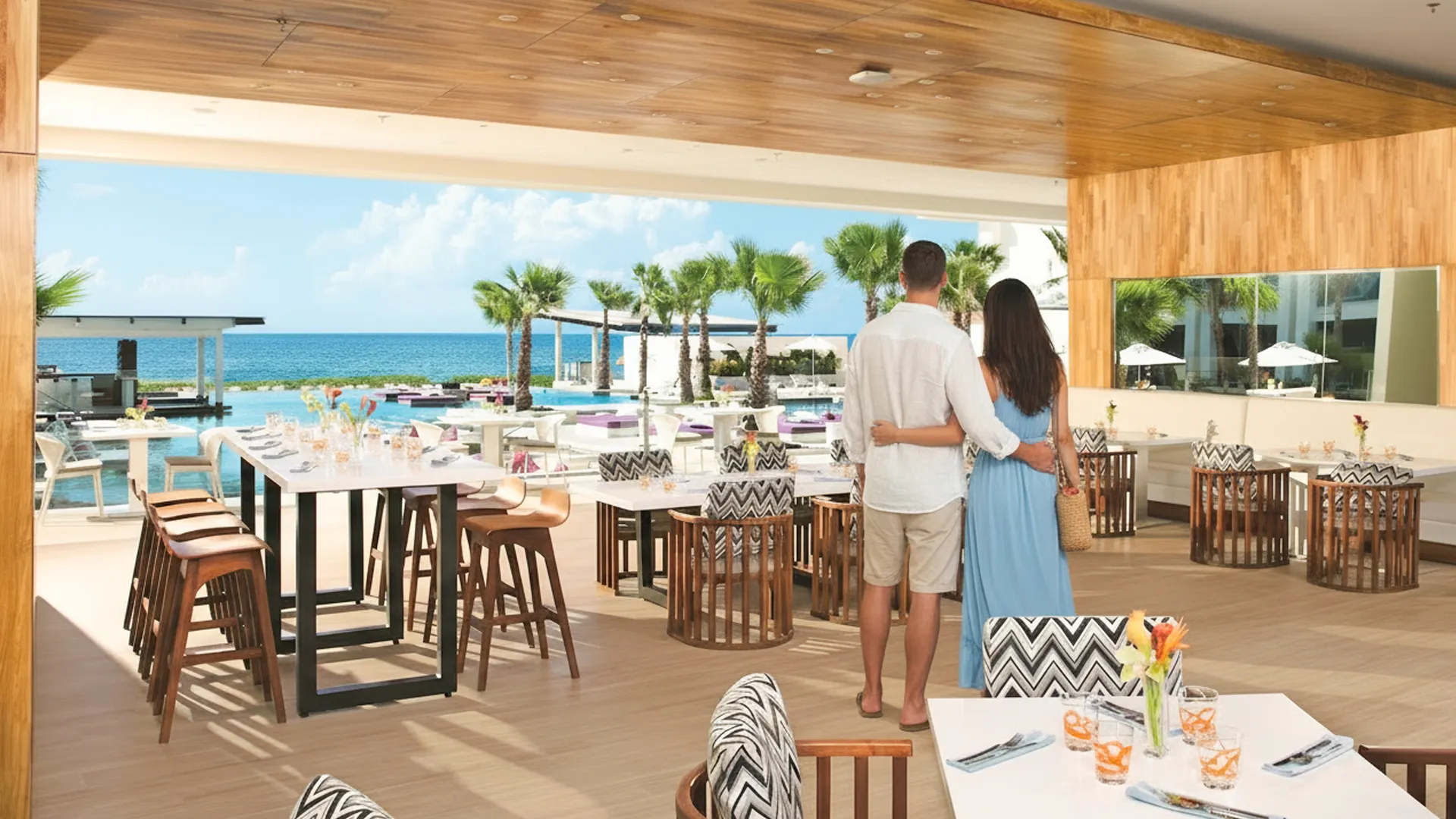 A couple standing arm in arm at an open-air restaurant in Breathless Cancun Soul, overlooking the turquoise ocean and palm trees.