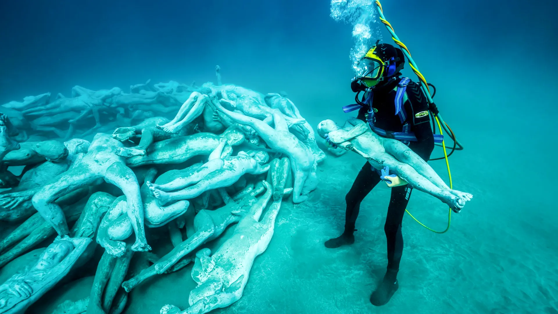 An underwater statue of a female figure with outstretched arms resembling wings, covered in marine growth, set against a blue ocean background.