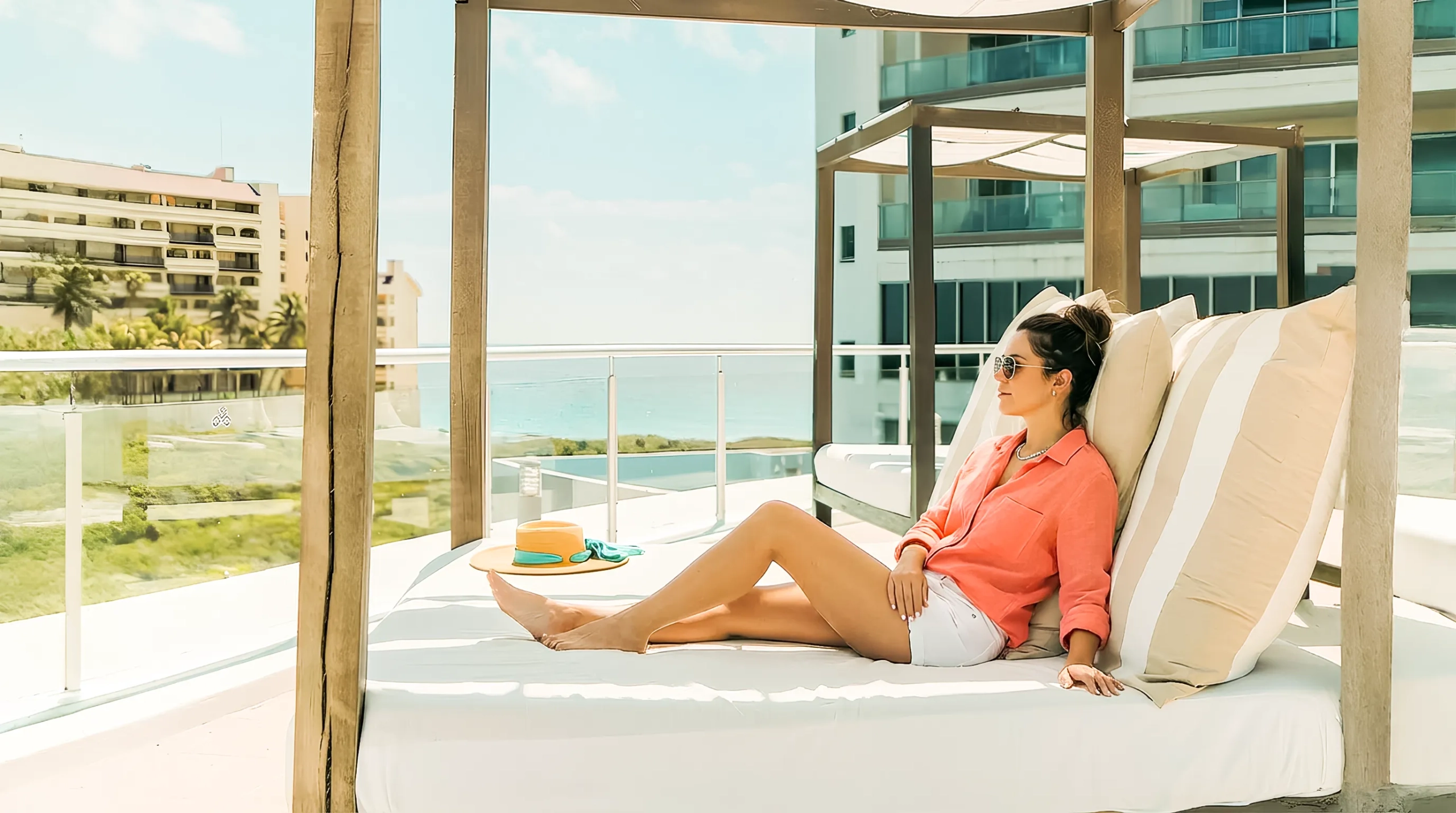Woman relaxing on a shaded cabana bed at Seadust Cancun, wearing sunglasses and sitting beside a sunhat with ocean views in the background.