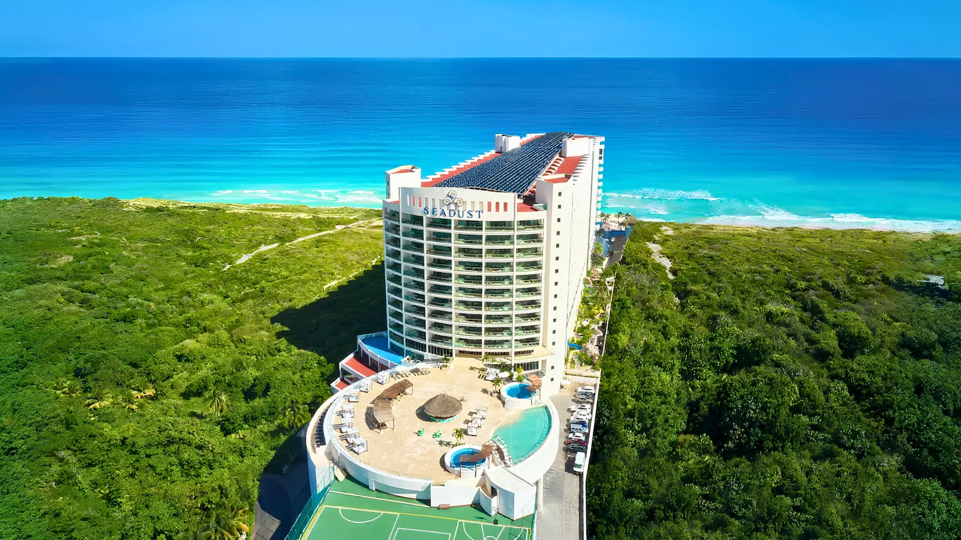 Aerial view of Seadust Cancun Family Resort surrounded by greenery, with a circular pool area, tall hotel tower, and bright turquoise ocean behind it.