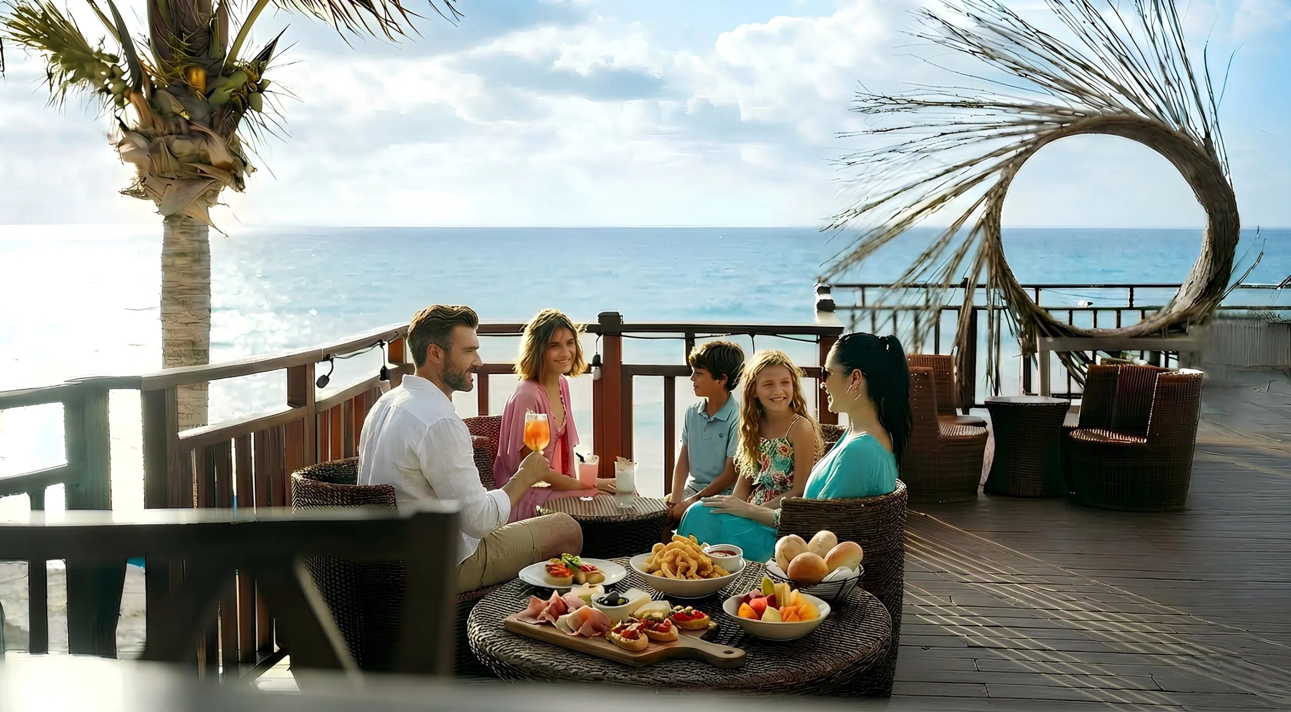 Family sitting together at an oceanfront deck at Seadust Cancun, enjoying drinks and snacks with a decorative palm-leaf arch in the background.