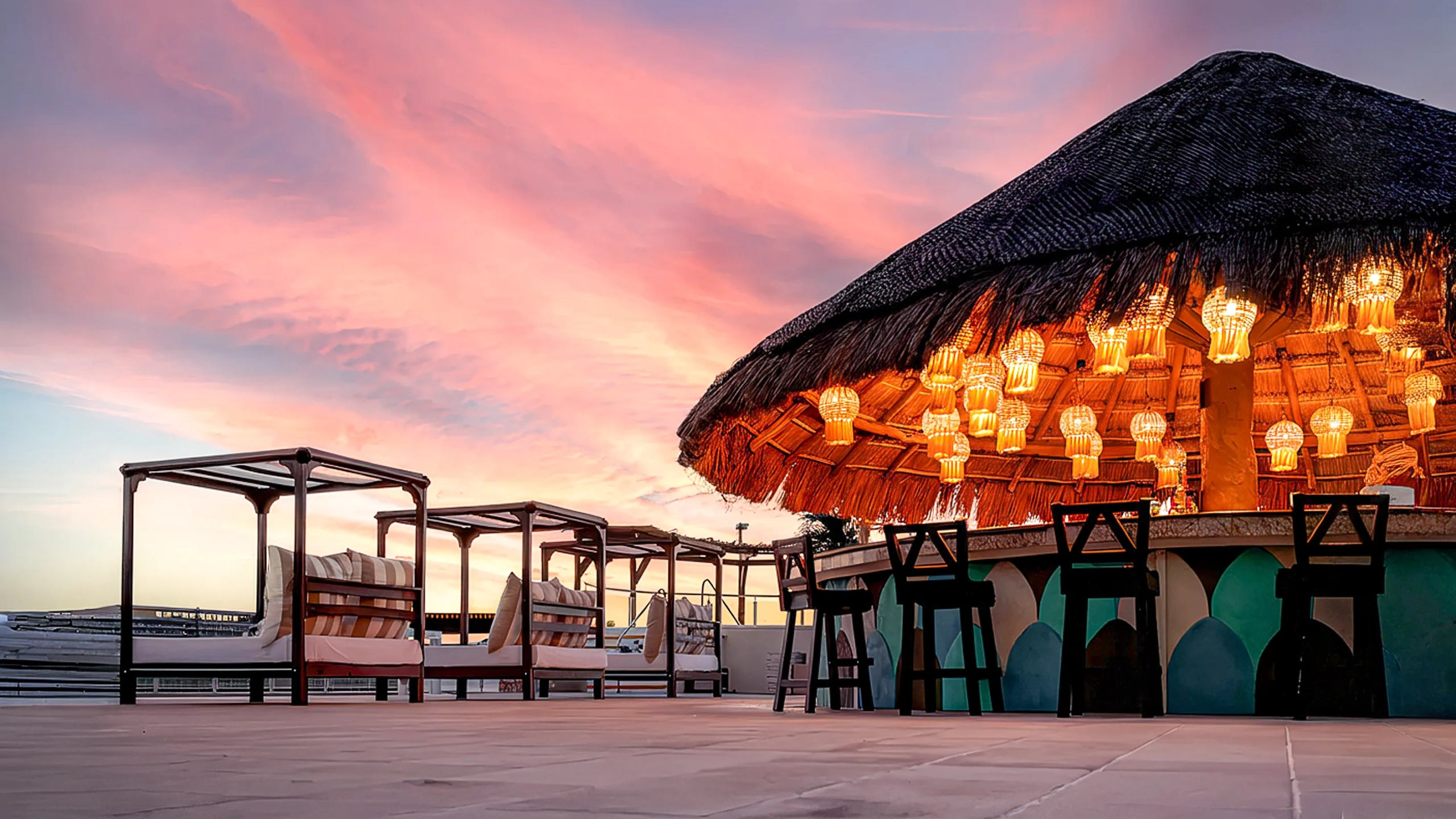 Outdoor beachfront bar at Seadust Cancun lit with warm hanging lanterns at sunset, next to canopy beds and lounge seating.