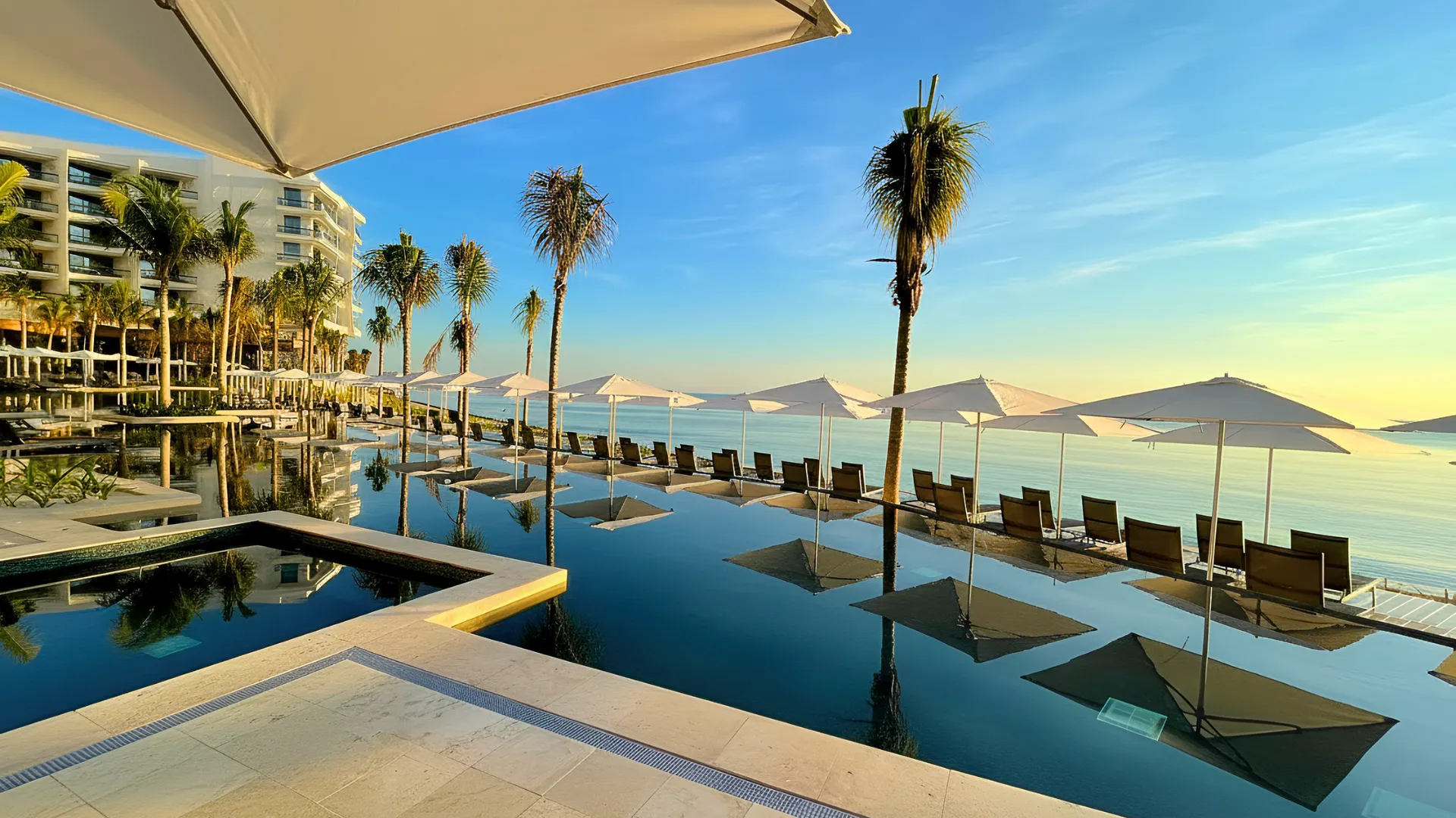 Infinity pool at Hilton Cancun All-Inclusive Resort surrounded by sun loungers and palm trees, overlooking the calm Caribbean Sea at sunrise.