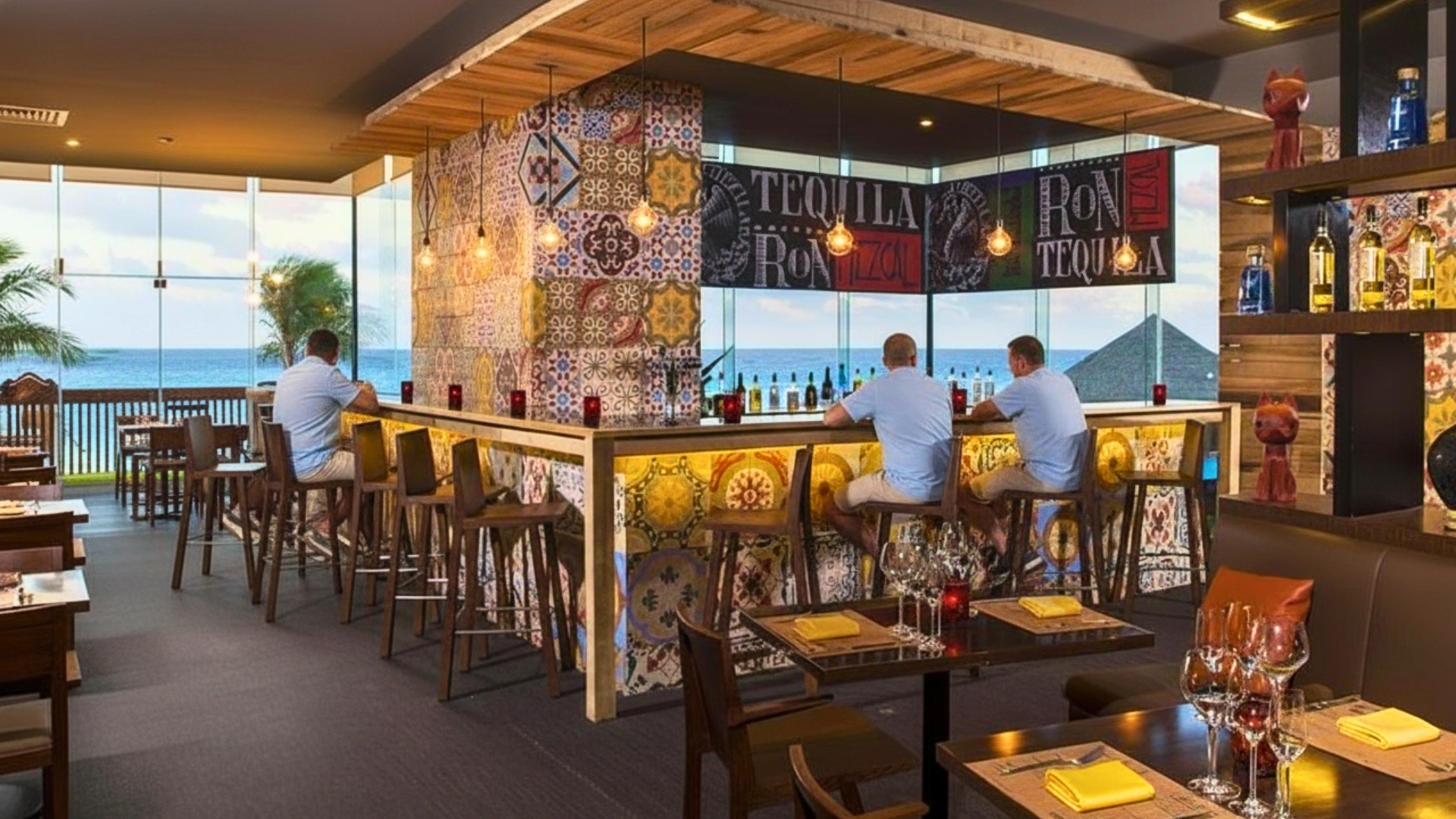 A vibrant restaurant with a tiled bar, wooden stools, and large windows overlooking the ocean, with guests seated and a decorative wall.