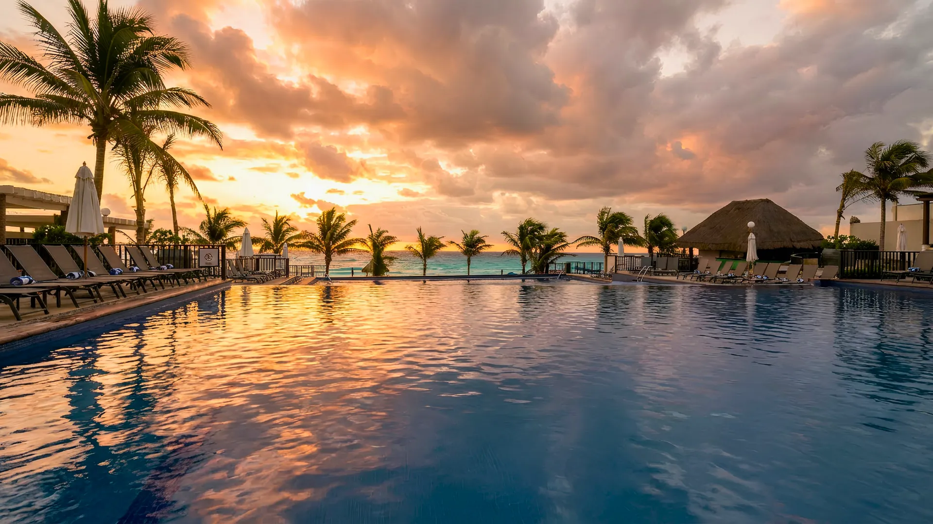 Sunset view over the main pool at Seadust Cancun with palm trees, loungers, and warm orange clouds reflecting on the water.