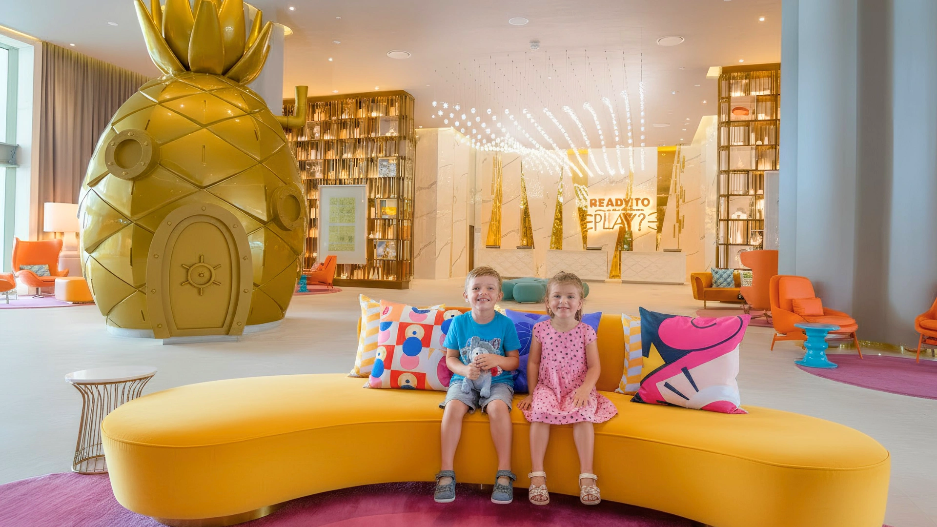 Two children sitting on a colorful sofa in the Nickelodeon Riviera Maya lobby, featuring SpongeBob’s pineapple decor.