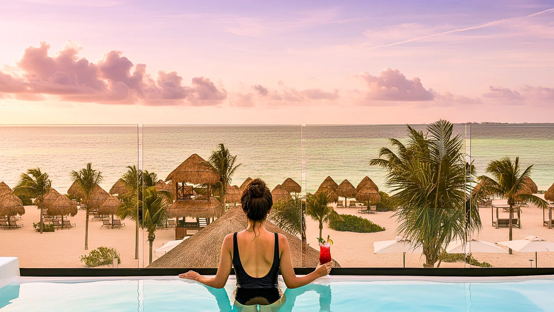  A person enjoying a sunset view from a swim-up suite in Excellence Riviera Cancun, overlooking the beach and resort area with a tropical drink in hand.