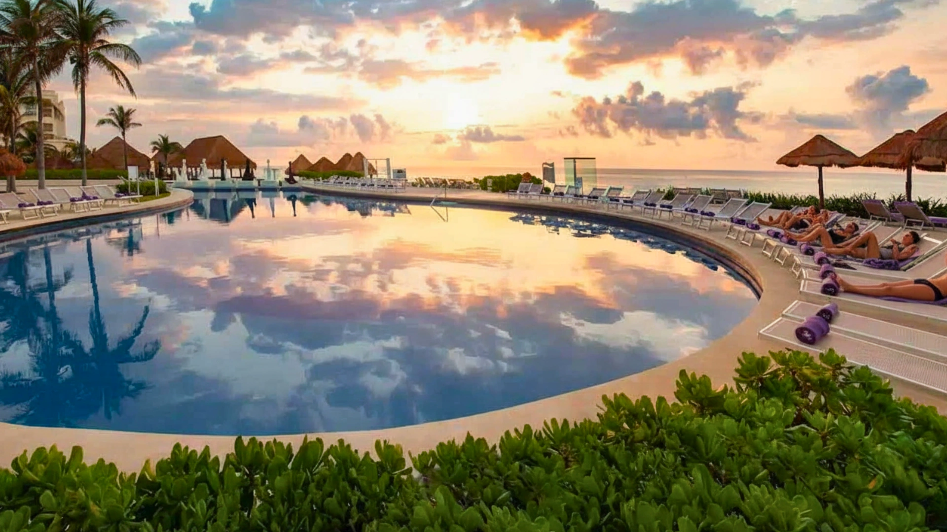 A large, curved swimming pool reflecting a sunset sky, surrounded by lounge chairs, thatched umbrellas, and palm trees.