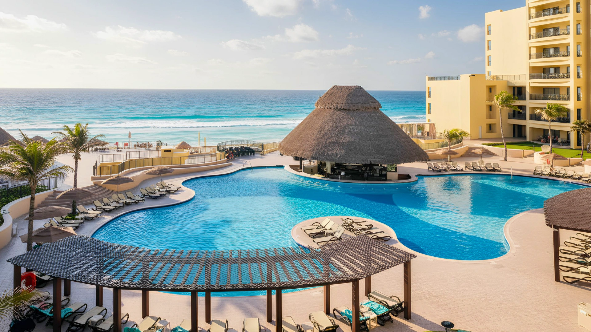 A shot of the resort pool area at Royal Sands Cancun with a thatched-roof bar and beach chairs.
