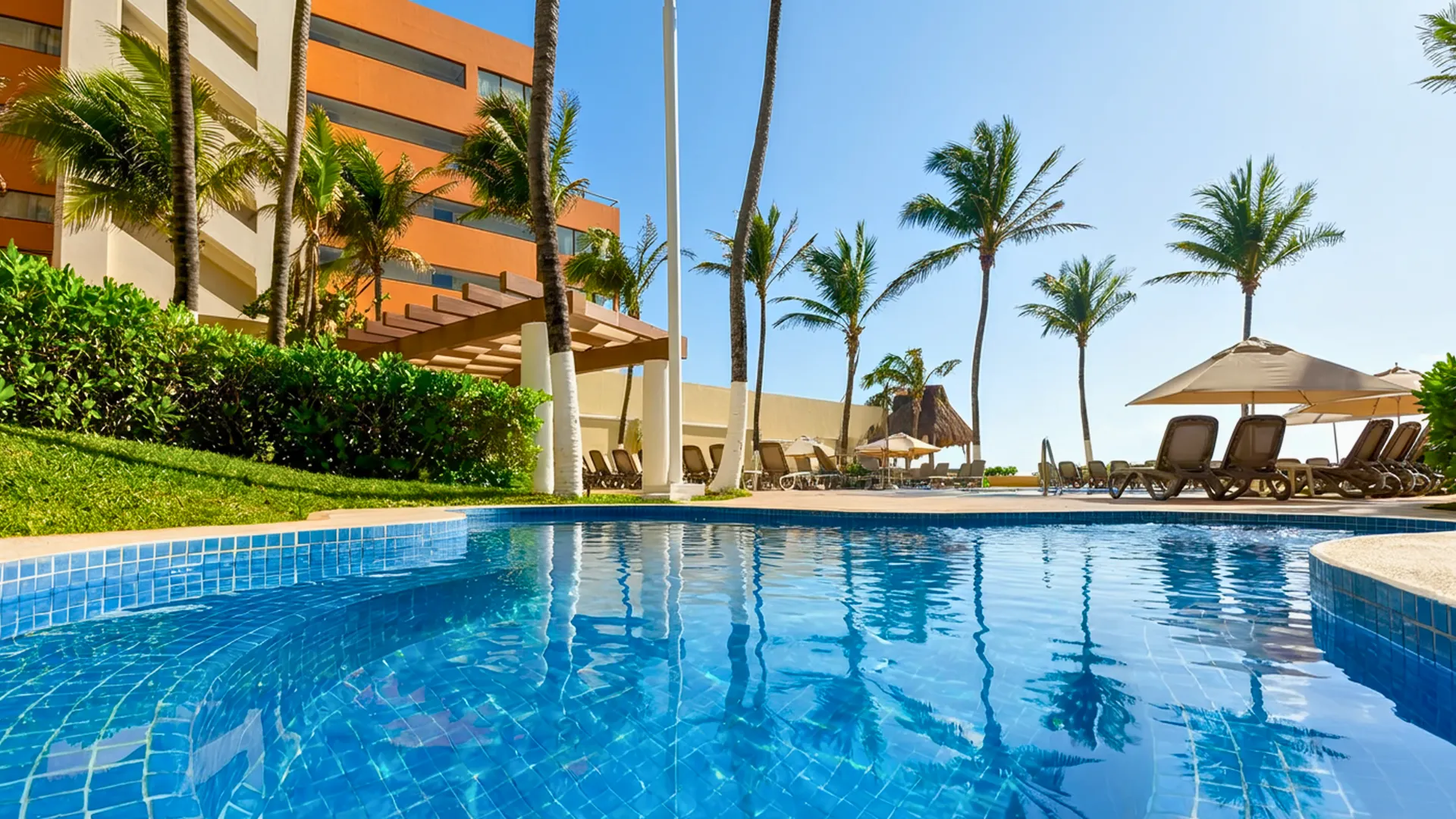 Outdoor pool at GR Solaris Cancun surrounded by palm trees, sun loungers, and bright blue sky.