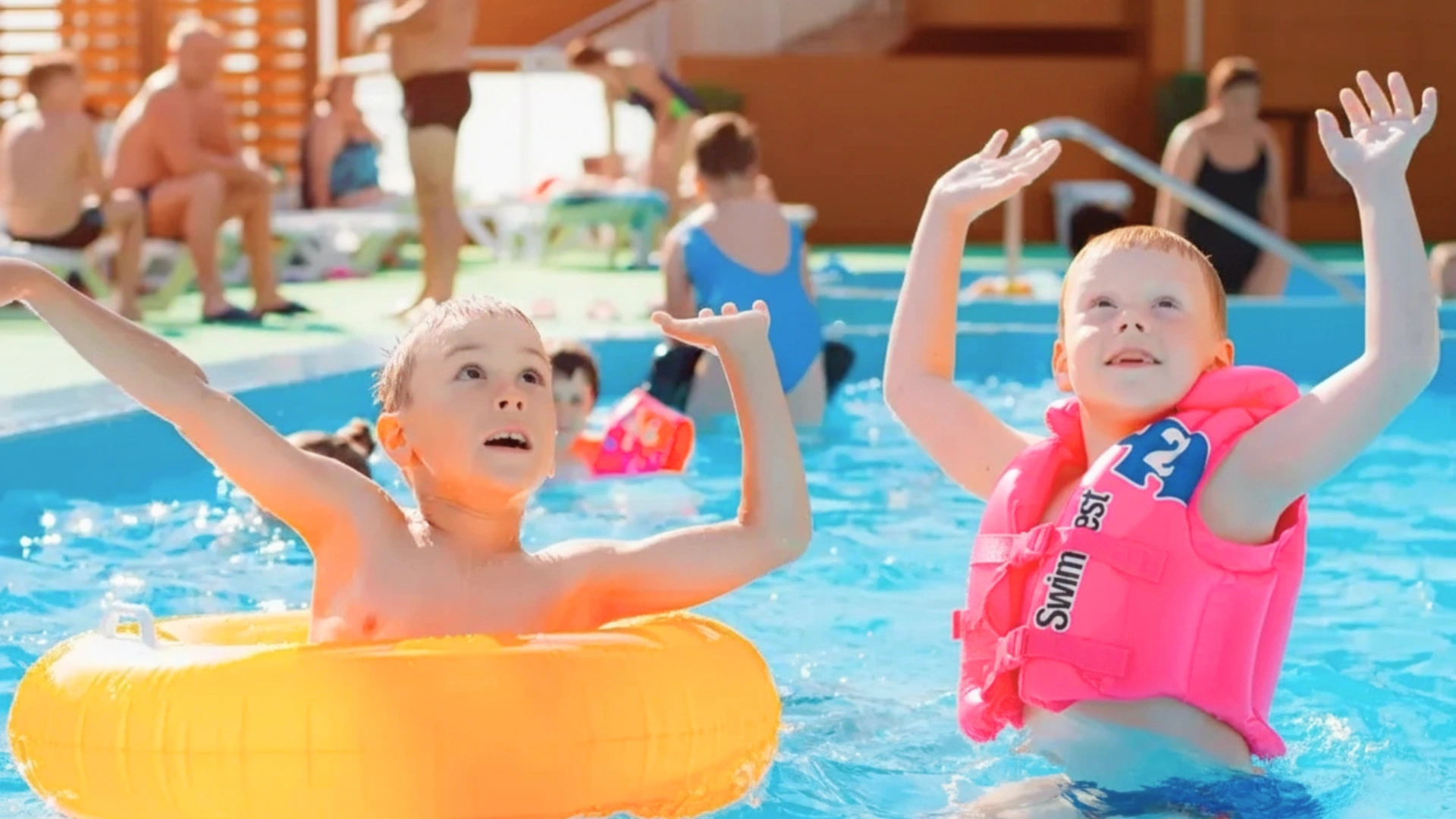 Children and adults enjoying a pool, with kids wearing floaties and raising their hands in excitement.