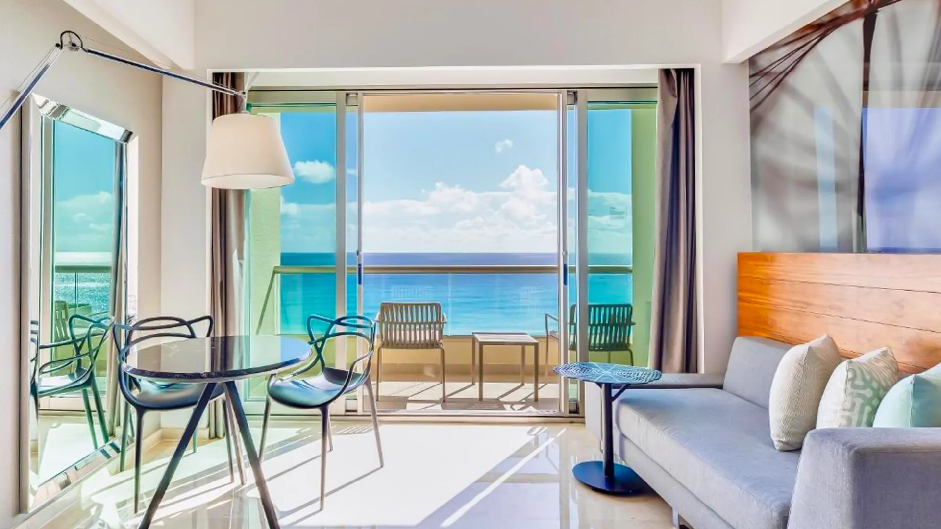 Spacious suite at Live Aqua Cancun with a gray sofa, round black dining table with green chairs, a side table with a lamp, large glass doors opening to a balcony with ocean views, and a wooden accent wall.
