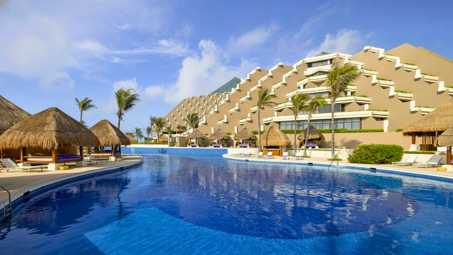  A luxurious resort with a large swimming pool, thatched-roof cabanas, and a multi-tiered building with palm trees under a clear blue sky.