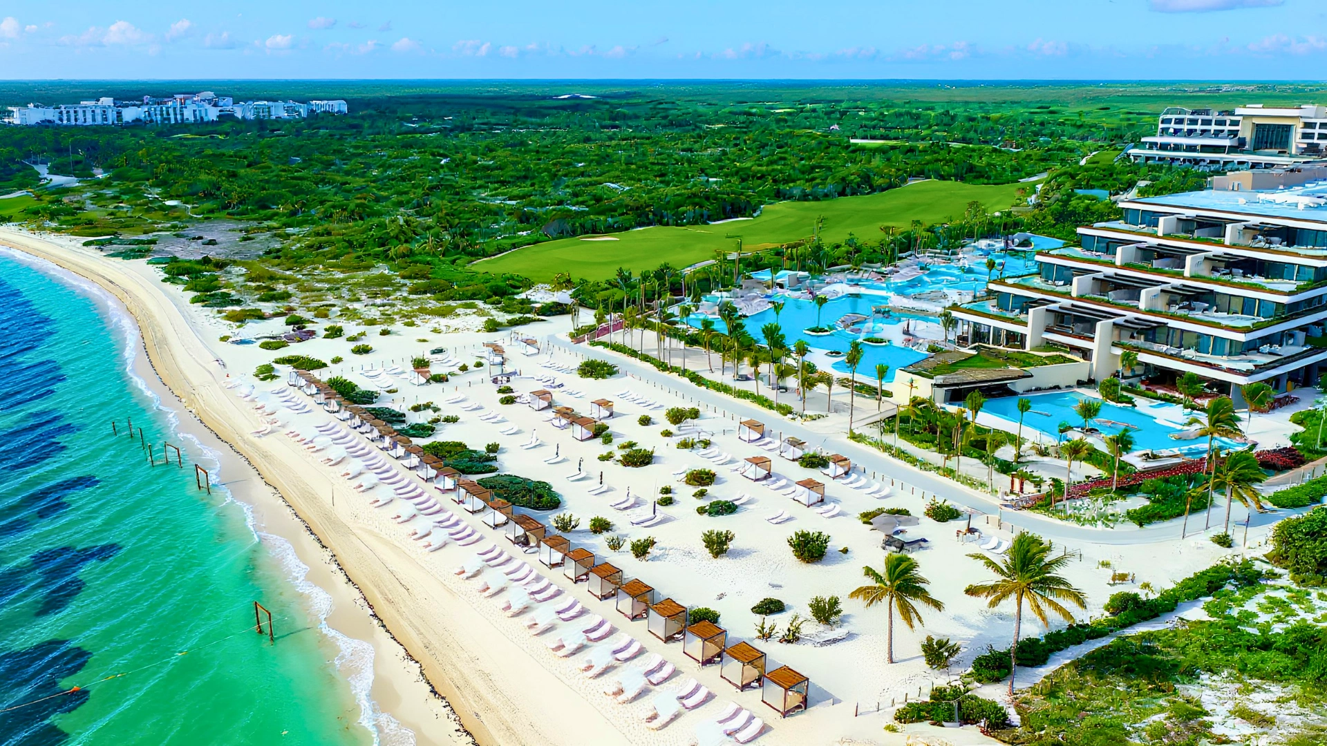 Aerial view of Atelier Playa Mujeres showcasing its beach, luxurious pool area, and vibrant palm trees. The image highlights the resort's expansive grounds and oceanfront location.